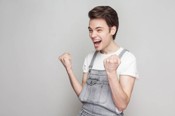 Portrait of happy amazed winner young brunette man in casual style with white t-shirt and denim overalls screaming and celebrating his victory on gray background - Fotografie, Obrázek