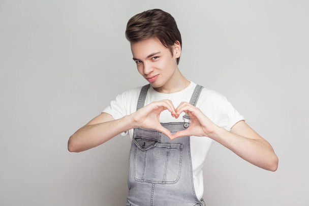Portrait of happy young brunette man in casual style with white t-shirt and denim overalls looking at camera and showing heart gesture on gray background - Valokuva, kuva