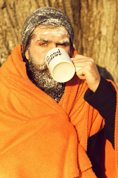 Frozen man, bearded hipster, with beard and moustache covered with white frost wrapped in orange blanket with warming drink in cup on winter day outdoors on natural backgroun - Foto, imagen