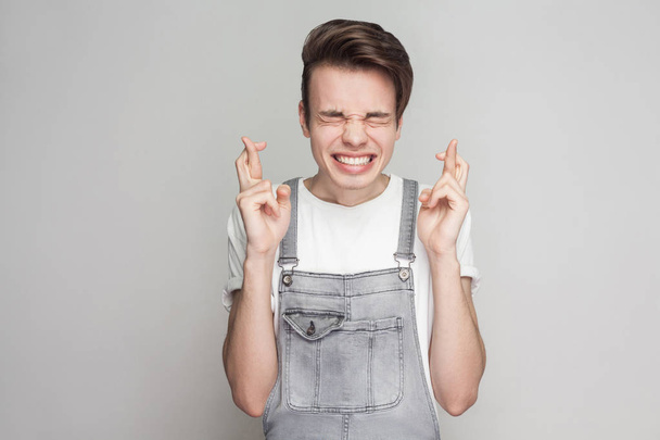 Portrait of hopeful young brunette man in casual style with white t-shirt and denim overalls standing with closed eyes and crossed fingers with clenched teeth on gray background - Foto, imagen
