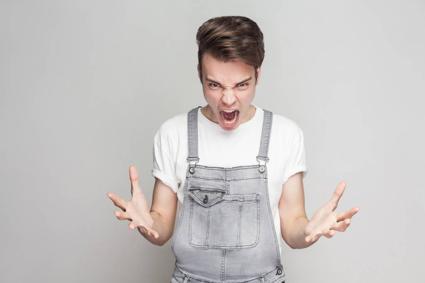 Portrait of angry young brunette man in casual style with t-shirt and denim overalls looking at camera with aggressive face and screaming on gray background - Foto, Imagem