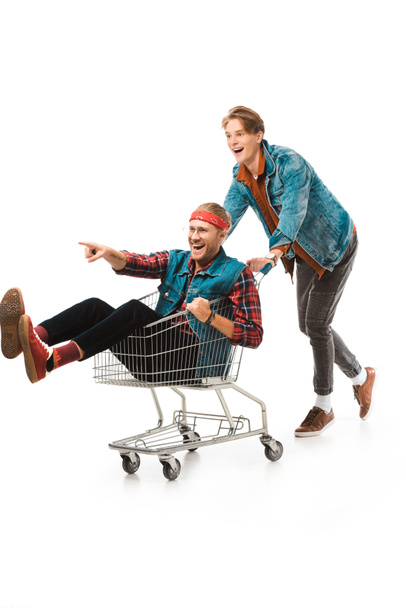 young hipster man carrying shopping cart with male friend pointing by finger isolated on white - Photo, Image