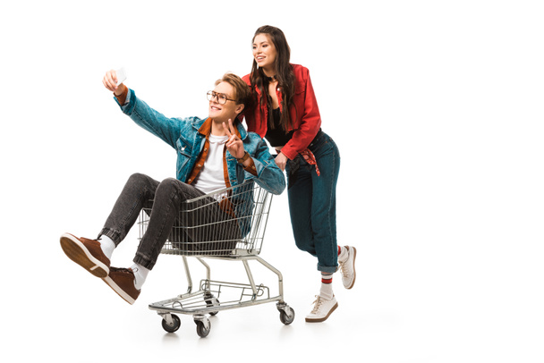 hipster masculino en carrito de compras haciendo señal de paz y tomando selfie mientras su novia de pie detrás aislado en blanco
 - Foto, imagen