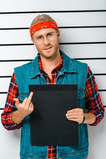 arrested hipster man in eyeglasses showing middle finger and holding empty prison board in front of police line up - Foto, imagen
