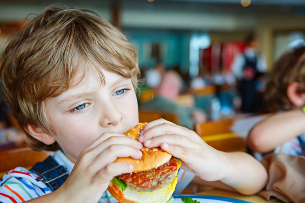Ładny chłopak przedszkola zjada hamburger siedzi w stołówce szkolnej - Zdjęcie, obraz