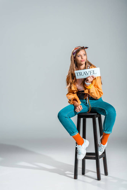stylish girl holding travel newspaper while sitting on stool on grey with copy space - Zdjęcie, obraz