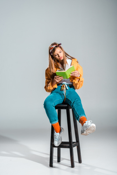 stylish student reading book while sitting on stool on grey - 写真・画像