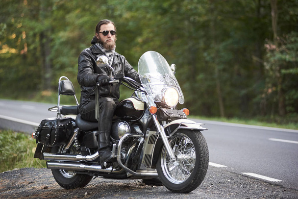 Handsome bearded motorcyclist in black leather clothing and dark sunglasses riding on motorcycle on country roadside on background of empty straight asphalt road and green trees bokeh foliage. - Photo, Image