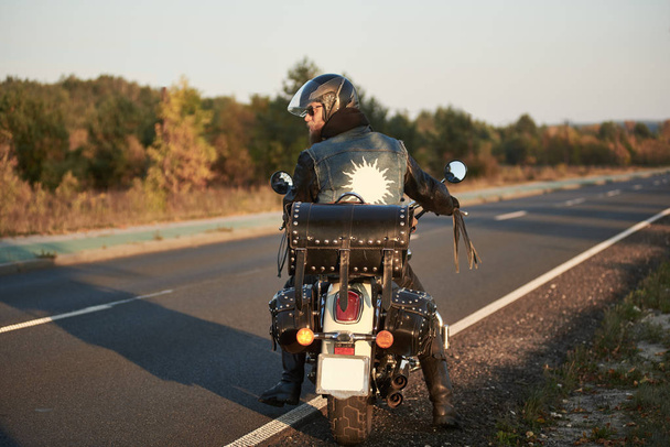 Zadní pohled na krásný biker v černé kožené bundě a sluneční brýle na motocykl cruiser na rozostřeného pozadí rovné silnice táhnoucí se k obzoru a zlaté podzimní stromy. - Fotografie, Obrázek