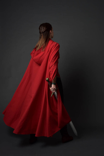Portret van de volledige lengte van brunette meisje dragen rode middeleeuws kostuum en mantel. staande poseren met terug naar de camera, houden een zwaard op grijs studio achtergrond. - Foto, afbeelding