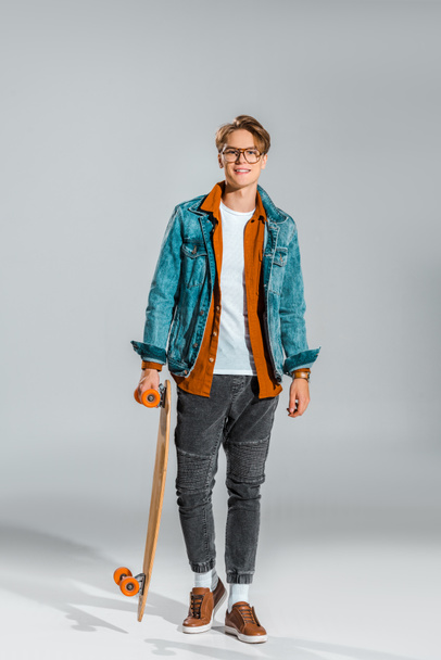 junger Hipster in Jeansjacke posiert mit Skateboard auf grau - Foto, Bild