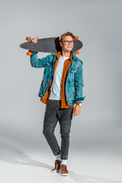 young male skateboarder in denim jacket posing with longboard on grey - Foto, imagen
