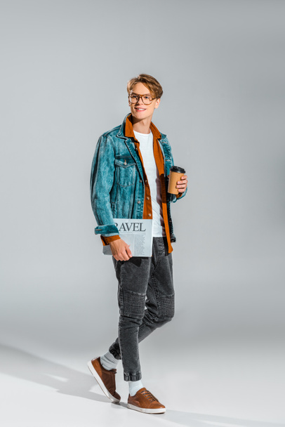 knappe hipster in denim jasje koffie houden om te gaan en reizen krant op grijs - Foto, afbeelding