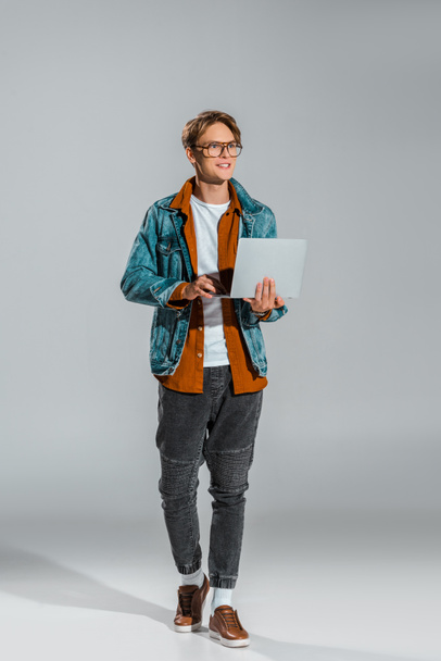handsome young freelancer in denim jacket using laptop on grey - Zdjęcie, obraz