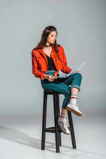 stylish young woman reading travel newspaper while sitting on stool on grey - Fotó, kép
