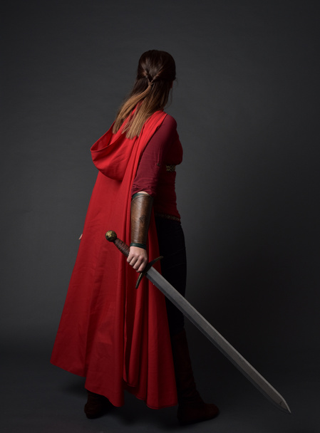Ganzkörperporträt eines brünetten Mädchens in rotem mittelalterlichen Kostüm und Mantel. mit dem Rücken zur Kamera stehend, ein Schwert auf grauem Studiohintergrund haltend. - Foto, Bild