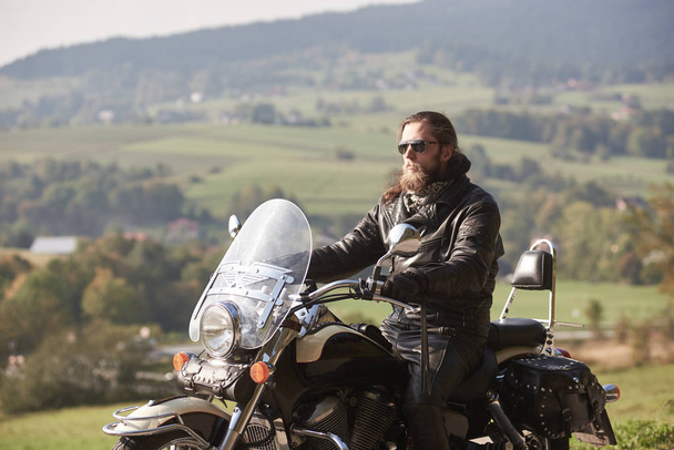 Handsome bearded rider with long hair in black leather jacket and sunglasses sitting on cruiser motorcycle on blurred background of green peaceful rural landscape and light foggy sky. - Photo, Image