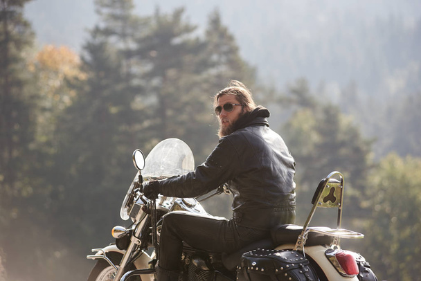 Hosszú haj fekete bőrkabát ül modern motorkerékpár elmosódott háttér lucfenyő magas fák és fás dombok a horizonton a szakállas motoros hátulnézete. - Fotó, kép