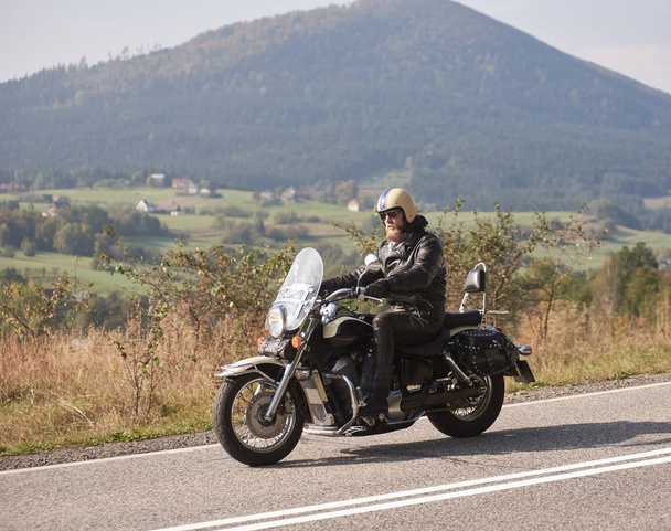 Vousatý motocyklista v přilbu, brýle a černé kožené oděvy jízda motocyklů mocný křižník se sunny asfaltové silnici na světlé letní den na pozadí zelené zalesněné kopce - Fotografie, Obrázek