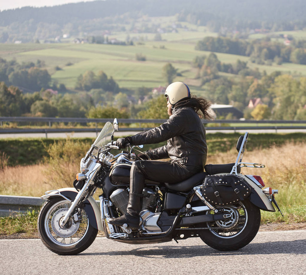 Kask ve siyah deri giyim binicilik bisiklet boyunca keskin sakallı motosikletçi yan görünüm parlak yaz günü kırsal manzara ve uzak yeşil tepeler puslu arka plan boş yol açmak. - Fotoğraf, Görsel