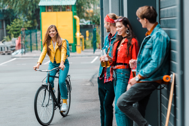 menina elegante atraente andar de bicicleta perto de amigos com garrafas de cerveja na rua
 - Foto, Imagem