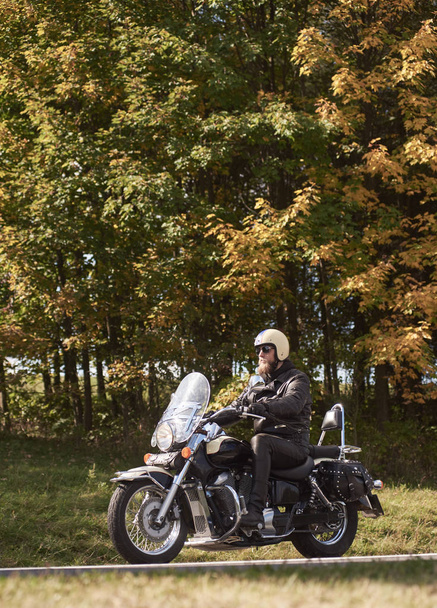 Vue latérale du motard barbu en casque, lunettes de soleil et vêtements en cuir noir à moto le long d'un étroit sentier asphalté par une journée ensoleillée d'été sur fond de grands arbres et d'herbe verte
. - Photo, image