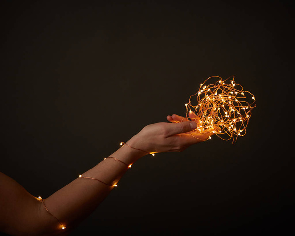 La mano de una mujer sostiene brillantes luces navideñas sobre un fondo negro con espacio para el texto. Diseño creativo de vacaciones
 - Foto, Imagen