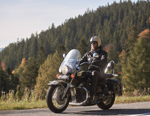 Handsome bearded biker in black leather jacket and sunglasses sitting on cruiser motorcycle on country roadside on blurred background of foggy green hills covered with dense spruce forest. - Photo, Image