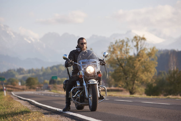 Portrait du beau motard barbu en veste en cuir noir sur moto de croisière sur le bord de la route de campagne sur fond flou de collines boisées vertes, de sommets de montagne blancs lointains et d'un ciel bleu vif
. - Photo, image