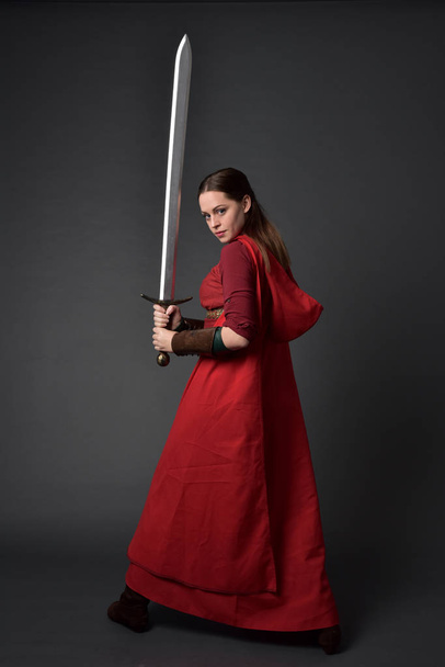 Pełna długość Portret brunetka dziewczyna ubrana w czerwony strój średniowiecznych i płaszcz. Stojący poza z powrotem do aparatu, trzymając miecz w studio szary tło. - Zdjęcie, obraz