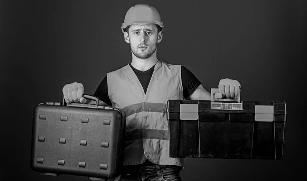 Worker, repairer, repairman, builder on thoughtful face choosing equipment for work. Man in helmet, hard hat holds toolbox and suitcase with tools, blue background. Toolbox and equipment concept - Foto, Imagem