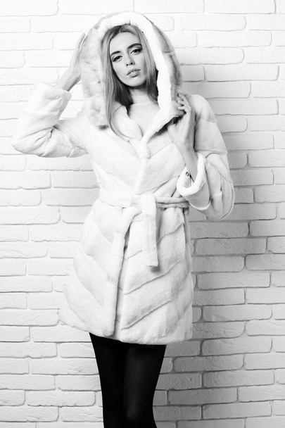 young fashionable sexy pretty woman or girl with beautiful long blonde hair in waist coat of white fur with hood and fashion makeup on brick wall studio background - Фото, зображення