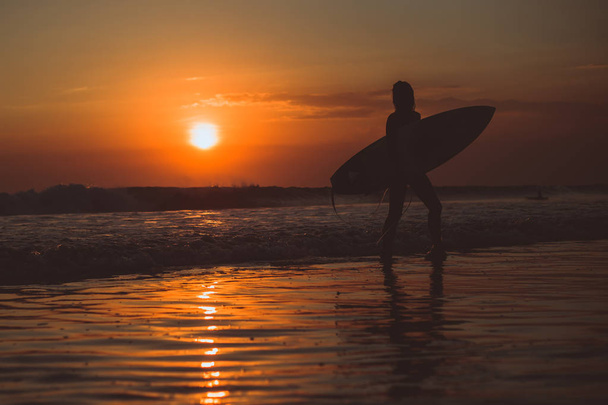 surfer woman walking with surfboard in sea water and enjoying sunset, orange sun in sky  - Photo, image