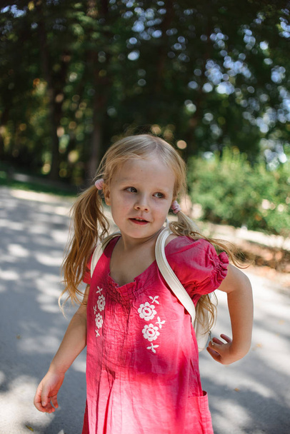 cute little blonde girl in red dress with white backpack walking at summer park  - Fotografie, Obrázek