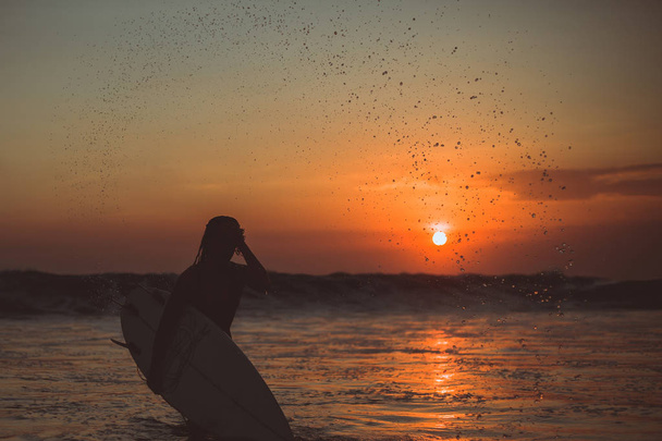 surfer woman silhouette with surfboard in sea, sunset reflection in water and splashes water drops  - Фото, изображение