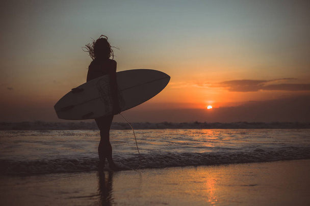 вид сзади на силуэт женщины, держащей доску для серфинга в морской воде на закате
  - Фото, изображение