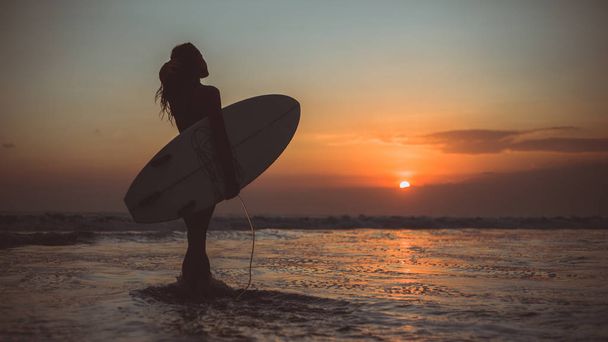 back view of woman silhouette holding surfboard in sea water at sunset  - Fotoğraf, Görsel