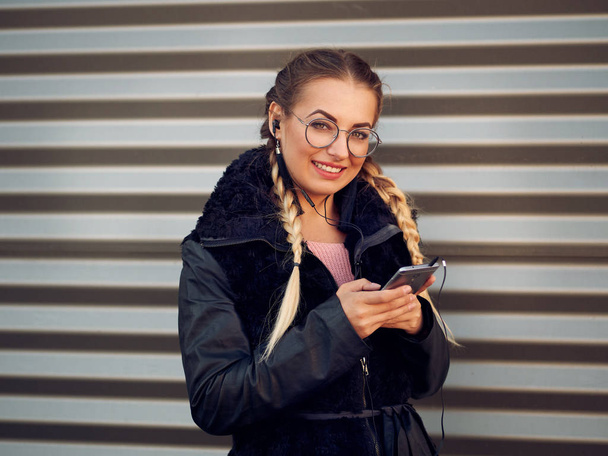portrait of a beautiful stylish young girl with braided blond hair in spikelets with round glasses in a black sheepskin coat with make-up listens to headphones from her phone on an iron striped background - Photo, Image