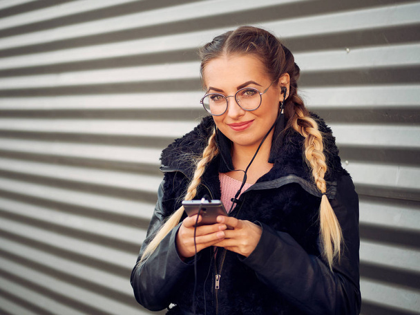 portrait of a beautiful stylish young girl with braided blond hair in spikelets with round glasses in a black sheepskin coat with make-up listens to headphones from her phone on an iron striped background - Photo, Image