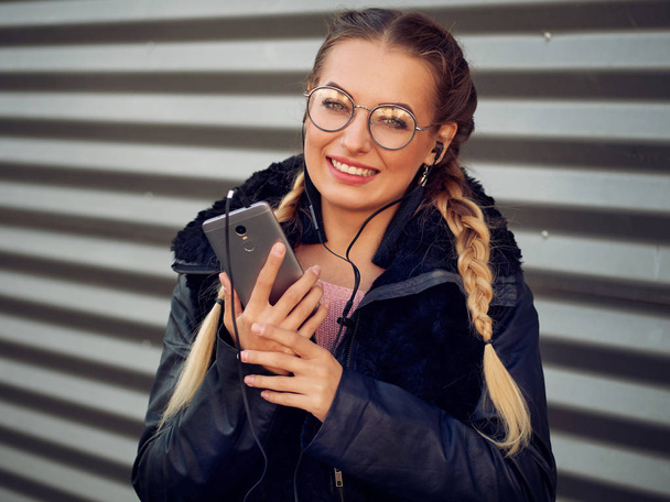 Portrait of a stylish young girl with braided blond hair in spikelets with round glasses with make-up listening to headphones in her phone music and drinking a hot drink on a striped iron background - Foto, afbeelding