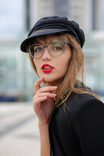 Gorgeous brunette woman with red lips wearing stylish cap and jacket, posing at the city - Фото, изображение