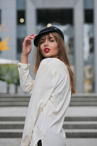 Stylish brunette woman wearing stylish cap and white shirt, posing near shopping mall - Fotó, kép