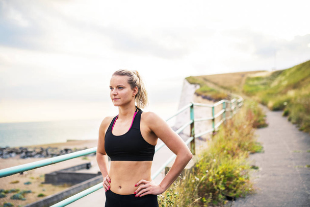 Young sporty woman runner in black activewear standing outside by the seaside, resting. - Foto, afbeelding