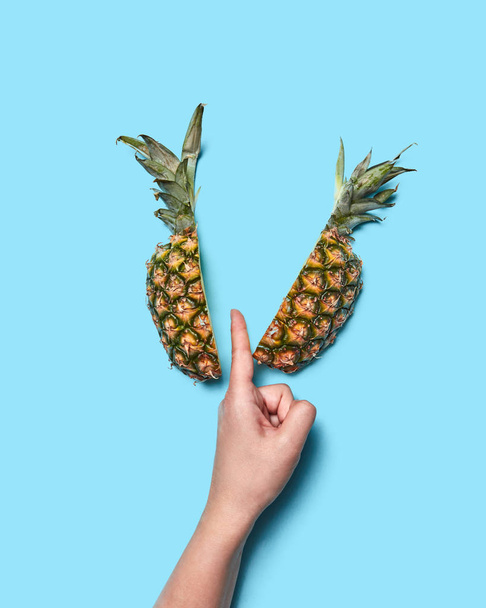 Female hand part with finger half of pineapple on blue background with space for text. Creative food composition. Flat lay - Фото, изображение