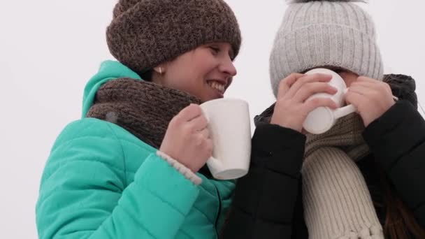 Beautiful girls drink coffee from white glass in winter park. Mom and adult daughter laugh and smile on a frosty day. - Footage, Video