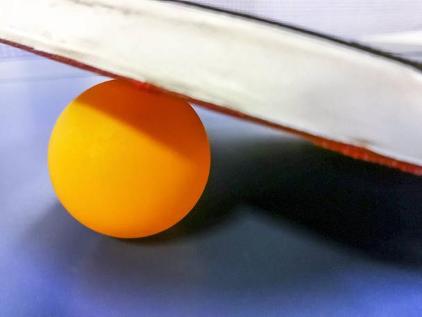 Raquete de tênis de mesa descansando em uma bola de laranja e uma mesa azul
 - Foto, Imagem