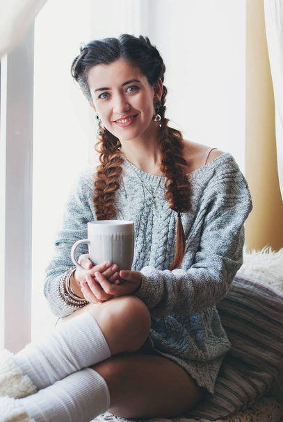 Young beautiful woman with mug of tea on the windowsill in the interior - sweet home concept - Фото, изображение