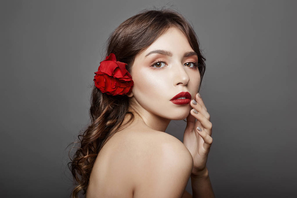 Woman with a big red flower in her hair. Brown-haired girl with a red flower posing on a gray background. Big beautiful eyes and natural makeup. Long curly hair, perfect face - Fotó, kép