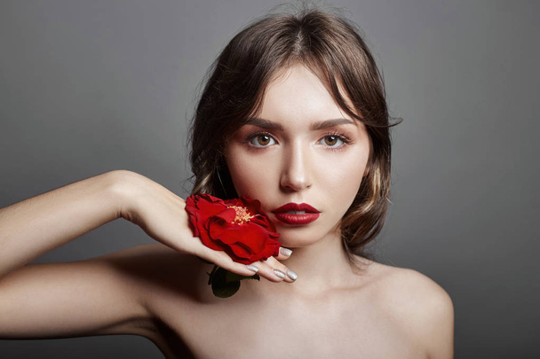 Woman with a big red flower in her hair. Brown-haired girl with a red flower posing on a gray background. Big beautiful eyes and natural makeup. Long curly hair, perfect face - Foto, Imagem