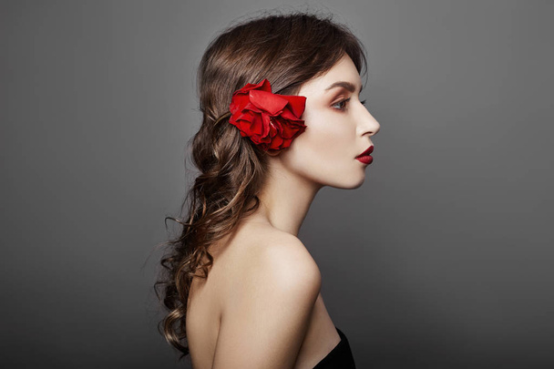 Γυναίκα με ένα μεγάλο κόκκινο λουλούδι στα μαλλιά της. Καστανά μαλλιά κορίτσι με ένα κόκκινο λουλούδι θέτοντας σε γκρι φόντο. Μεγάλα όμορφα μάτια και φυσικό μακιγιάζ. Μακριά σγουρά μαλλιά, τέλειο πρόσωπο - Φωτογραφία, εικόνα